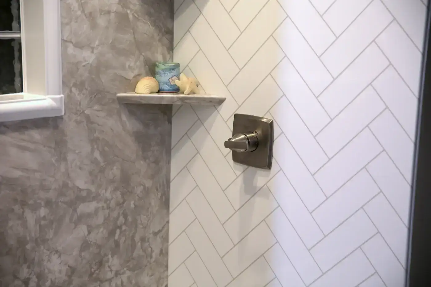 Custom bathtub wall surrounds combine white herringbone tile and grey marble panels with an integrated corner shelf and sleek metallic valve fixture.