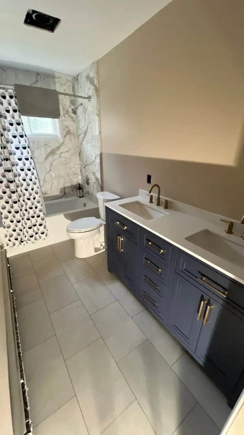 A stylish full bathroom remodel features a double-sink navy vanity with gold hardware, grey floor tiles, and a tub-shower combination surrounded by marble-patterned walls.