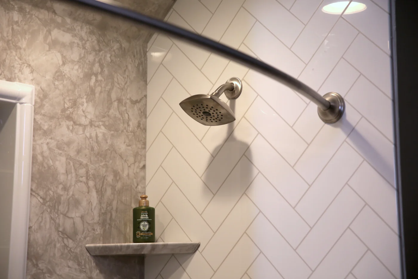 A brushed nickel showerhead is mounted against stylish bathtub wall surrounds featuring white herringbone tiles paired with a marbled accent panel.