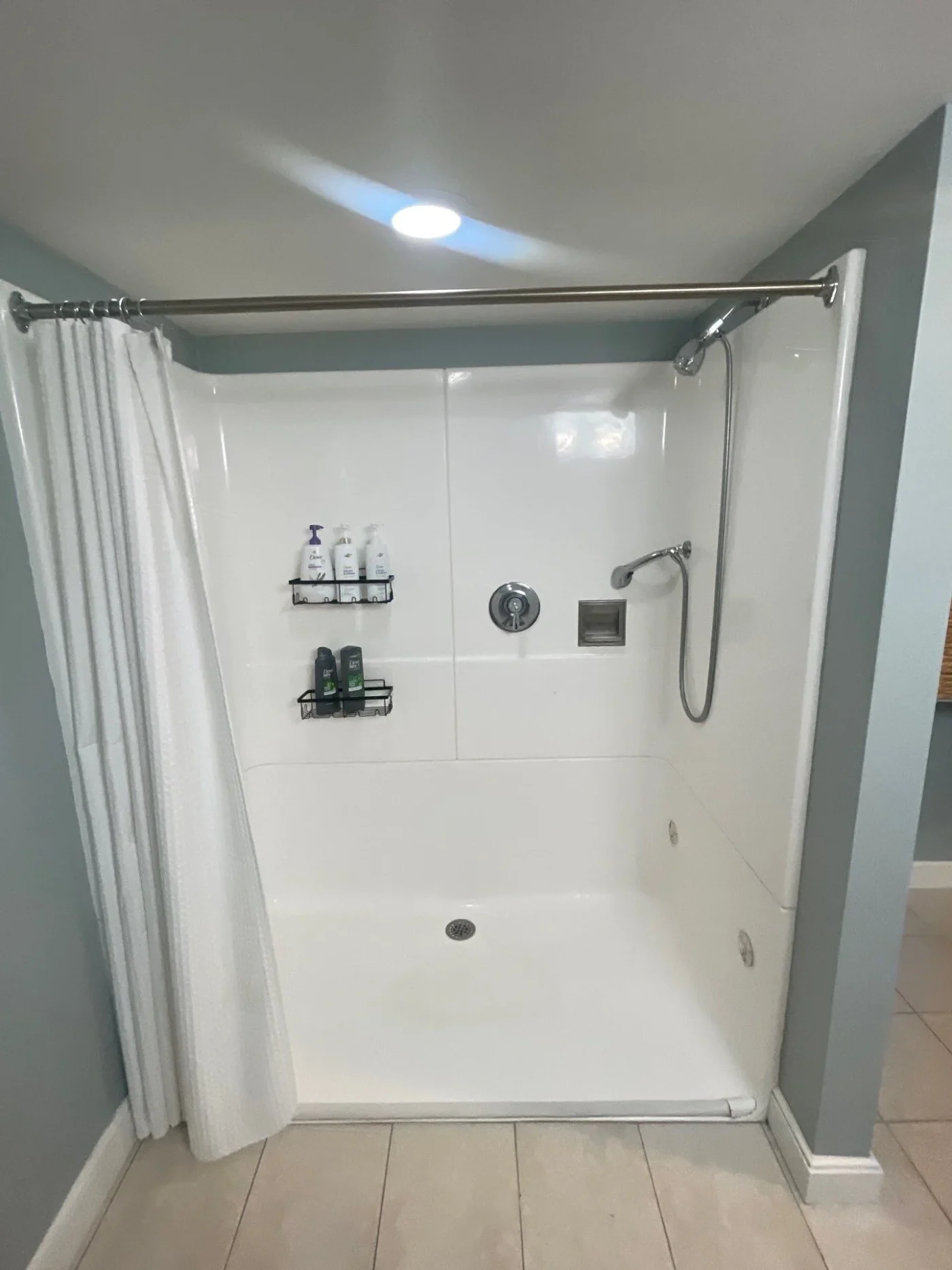 A clean, bright white shower base & wall installation features smooth surrounds, convenient shower caddies, and a handheld showerhead paired with a white curtain.