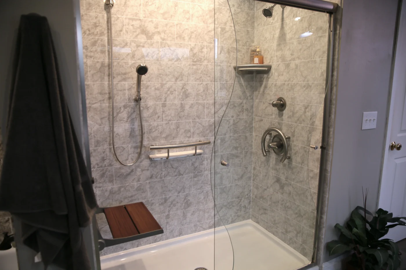 A modern walk-in shower with clear glass doors showcases a sleek shower base and wall installation featuring grey pattern surround, a fold-down wooden seat, and brushed nickel fixtures.