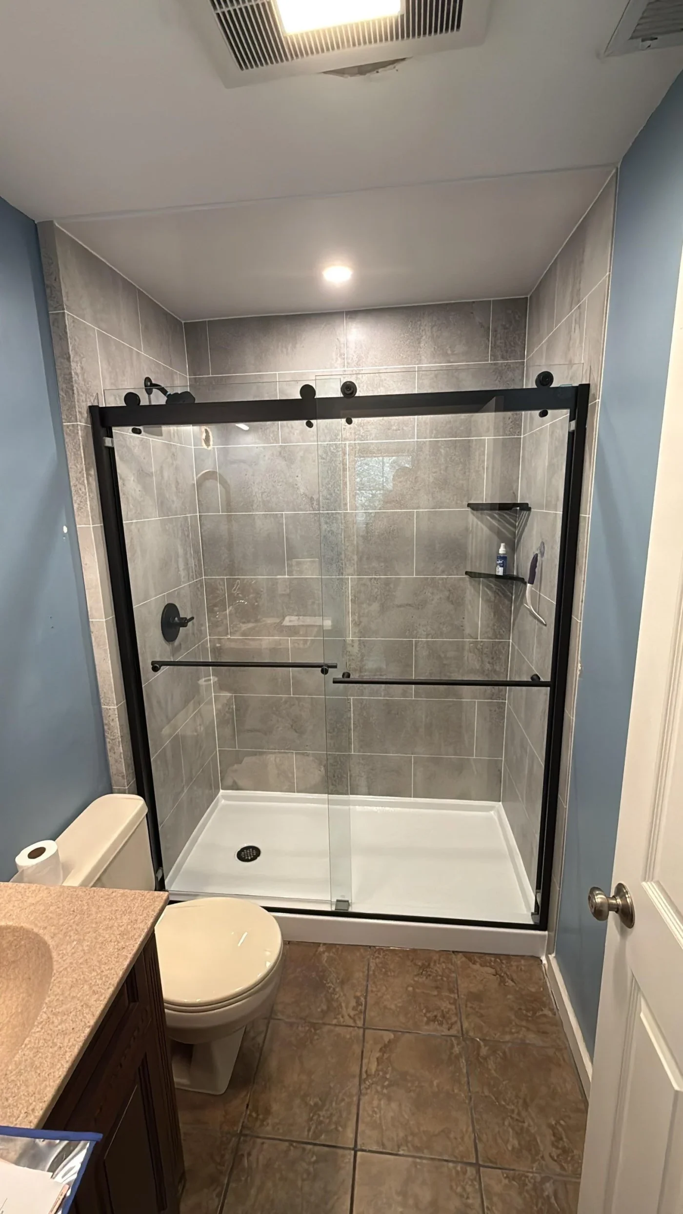A modern walk-in shower highlights a complete shower base & wall installation featuring grey stone-look tile surrounds and sliding glass doors with matte black hardware.