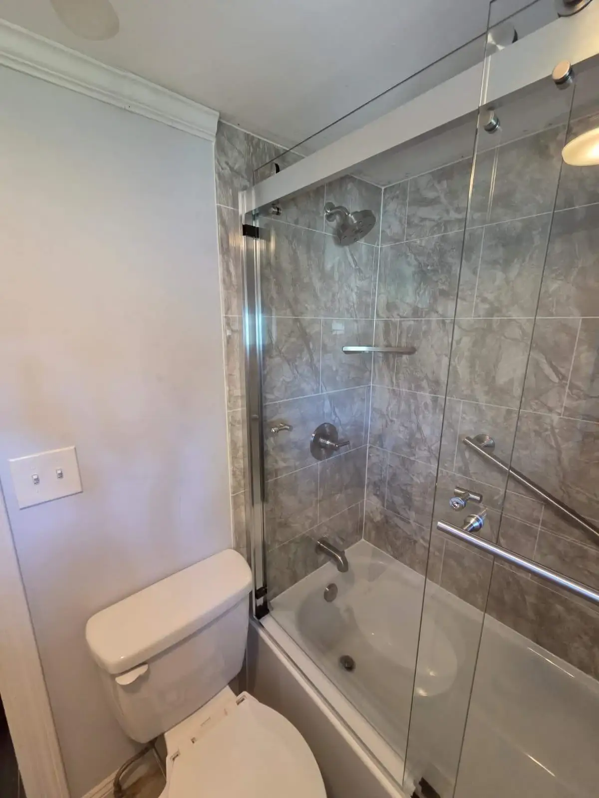 A stylish bathtub and shower combination features clear sliding glass doors paired with grey marble-patterned bathtub wall surrounds and modern brushed fixtures.