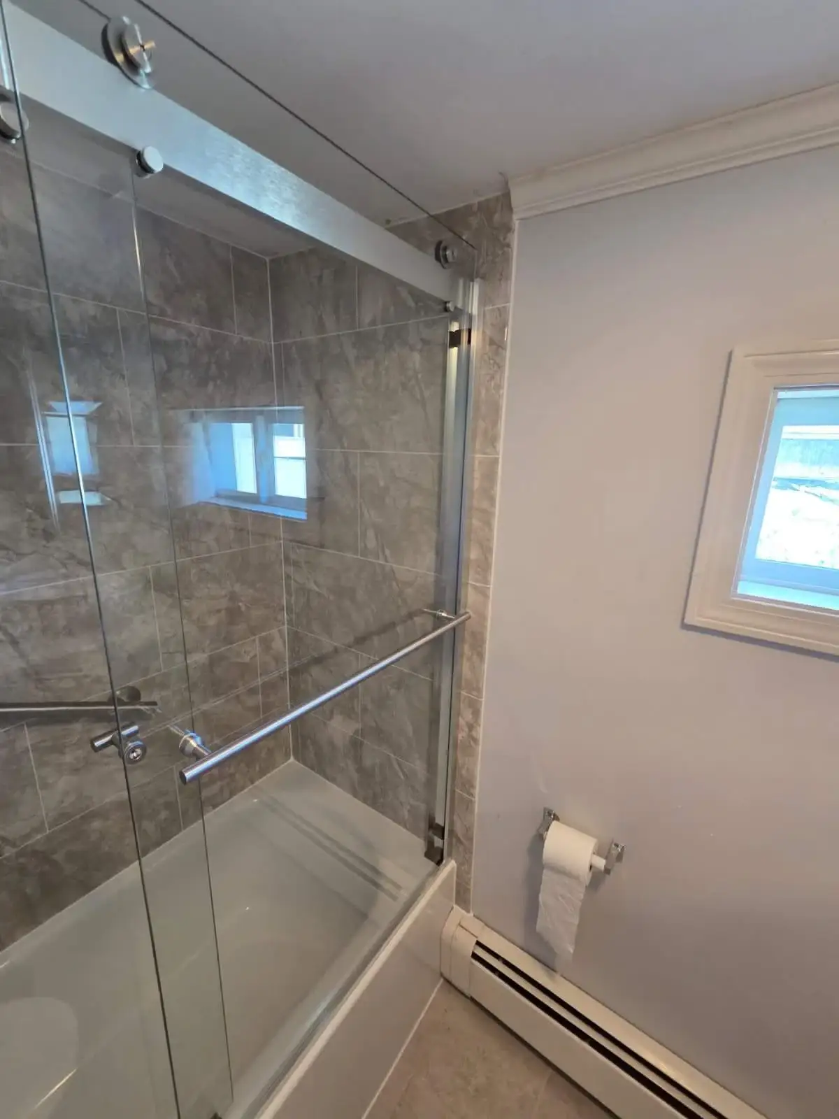 A modern bathtub and shower combination showcases clear sliding glass doors set against grey stone-patterned tile bathtub wall surrounds.